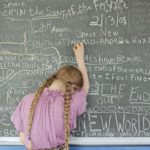 A young girl in pink writes on a blackboard.