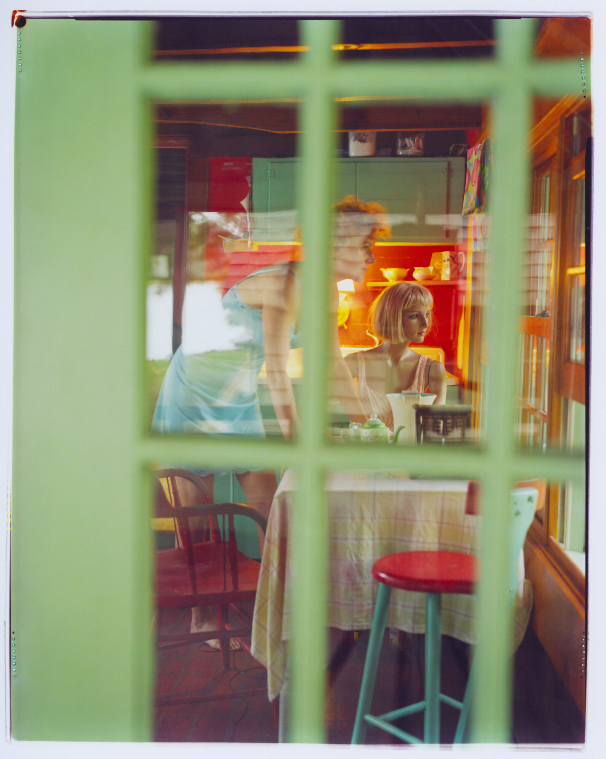 A woman and a mannequin through a green door window in a kitchen.