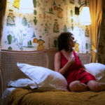 A woman in a red dress on bed with pillows.