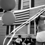 people watch the 4th of July parade with steamers and balloons and an American flag tagged to a building.