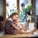 Young girl holding a doll with a boy looking in the window at her