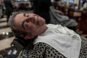 A man leans back on a chair at a salon. He is wearing a cheetah print cover and a hand towel on his chest.