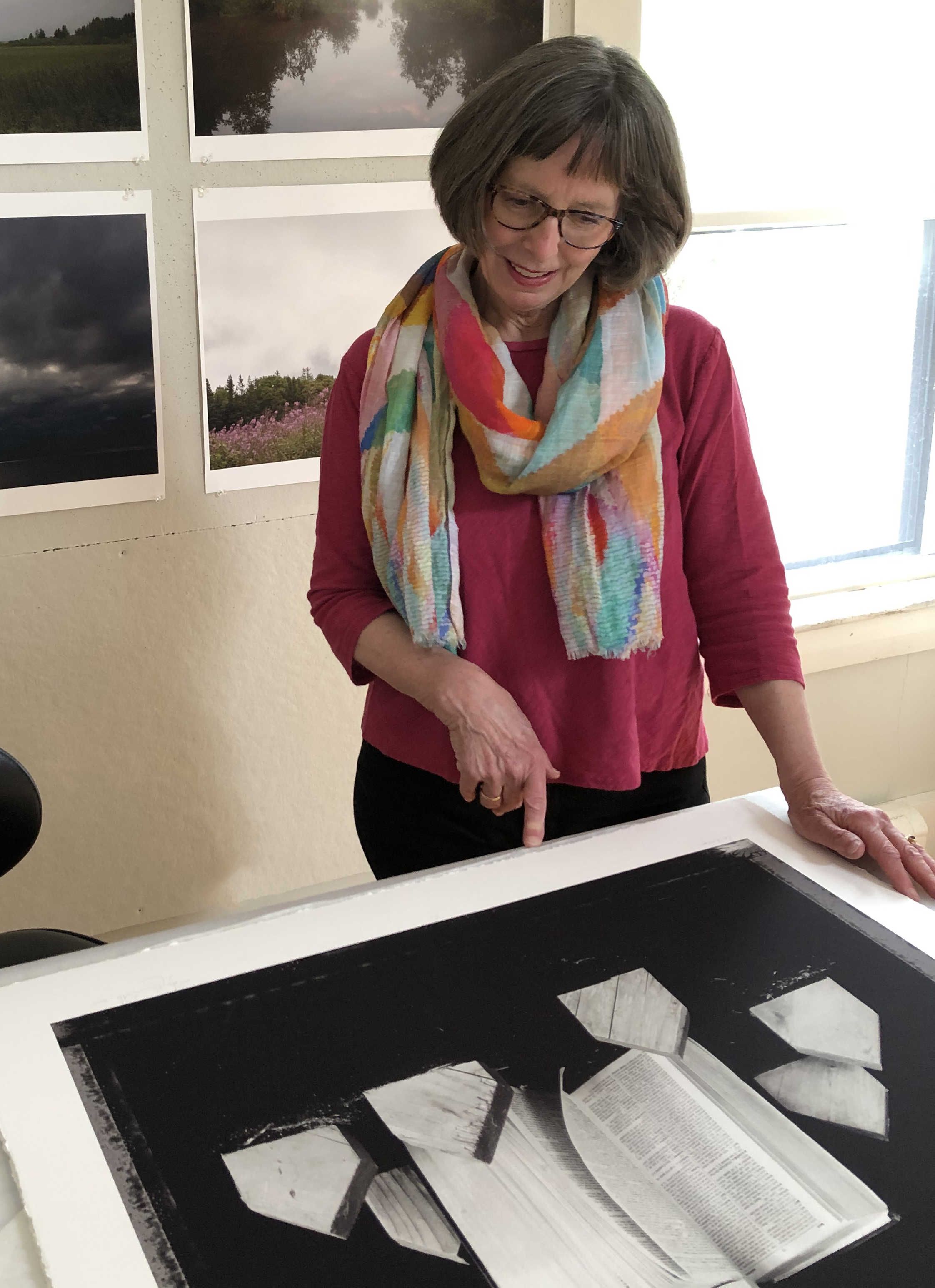 A woman with short hair wearing a scarf points at a photograph laying on a table. There is a grid of landscape photographs pinned to the back wall.