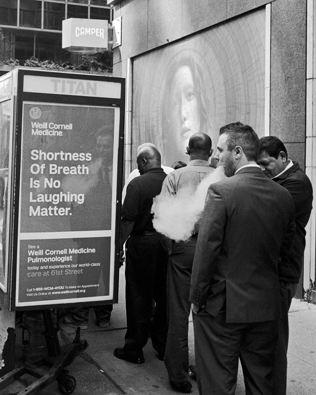 There are people in line to get into something. On the left is a sign about shortness of breath. A man standing beside the sign is smoking and blowing smoke.