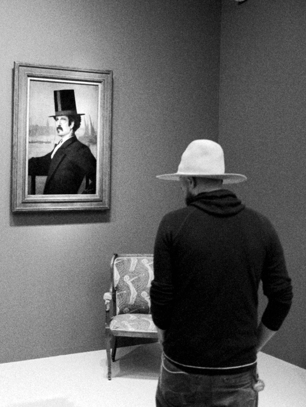 In a museum a man in a dark shirt wears a white cowboy hat. He looks at a painting of a man dressed formally and wears a top hat.