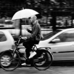 It is raining and a man on bicycle rides in the street with a white umbrella. He passes parked cars. As he is moving he is slightly blurred.