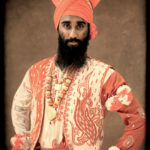 A man from India is dress in a white and orange decorated shirt and wears an orange turban.