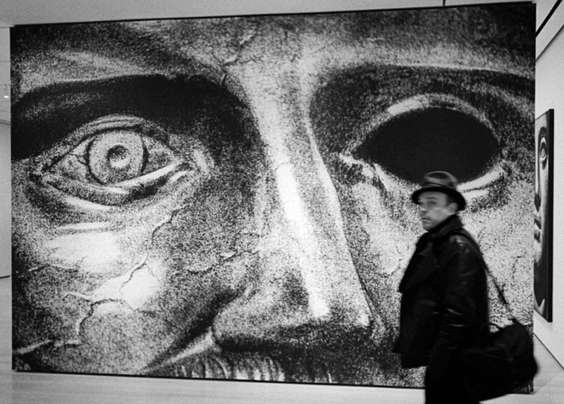 A large statue head photo is a wall decoration. The eyes and nose are featured only. A Mann dressed in black walks by with a valise over his shoulder.