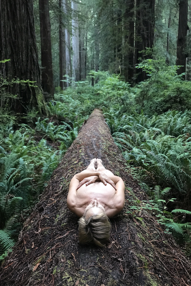 A nude woman lies on a fallen tree.