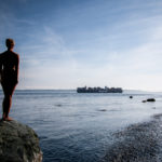 A nude woman stands on the shore and looks at an island.