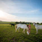 A nude woman is standing with horses.