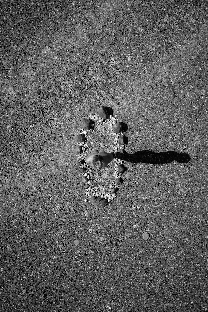 A nude woman is surrounded by stones in a circle. She is standing and casts a shadow.