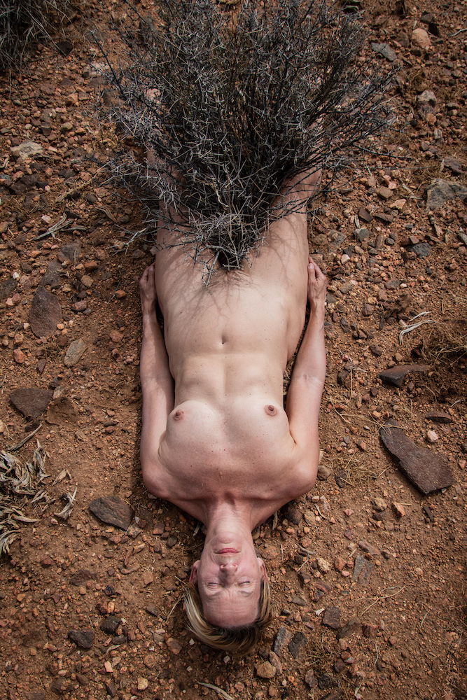 A nude woman lies on the ground and straddles a plant.