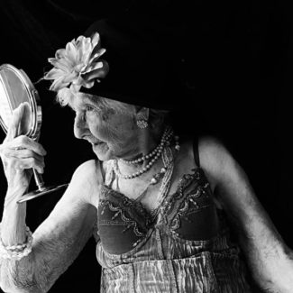 An older woman dressed in lingerie, head dress and jewels looks into a mirror .