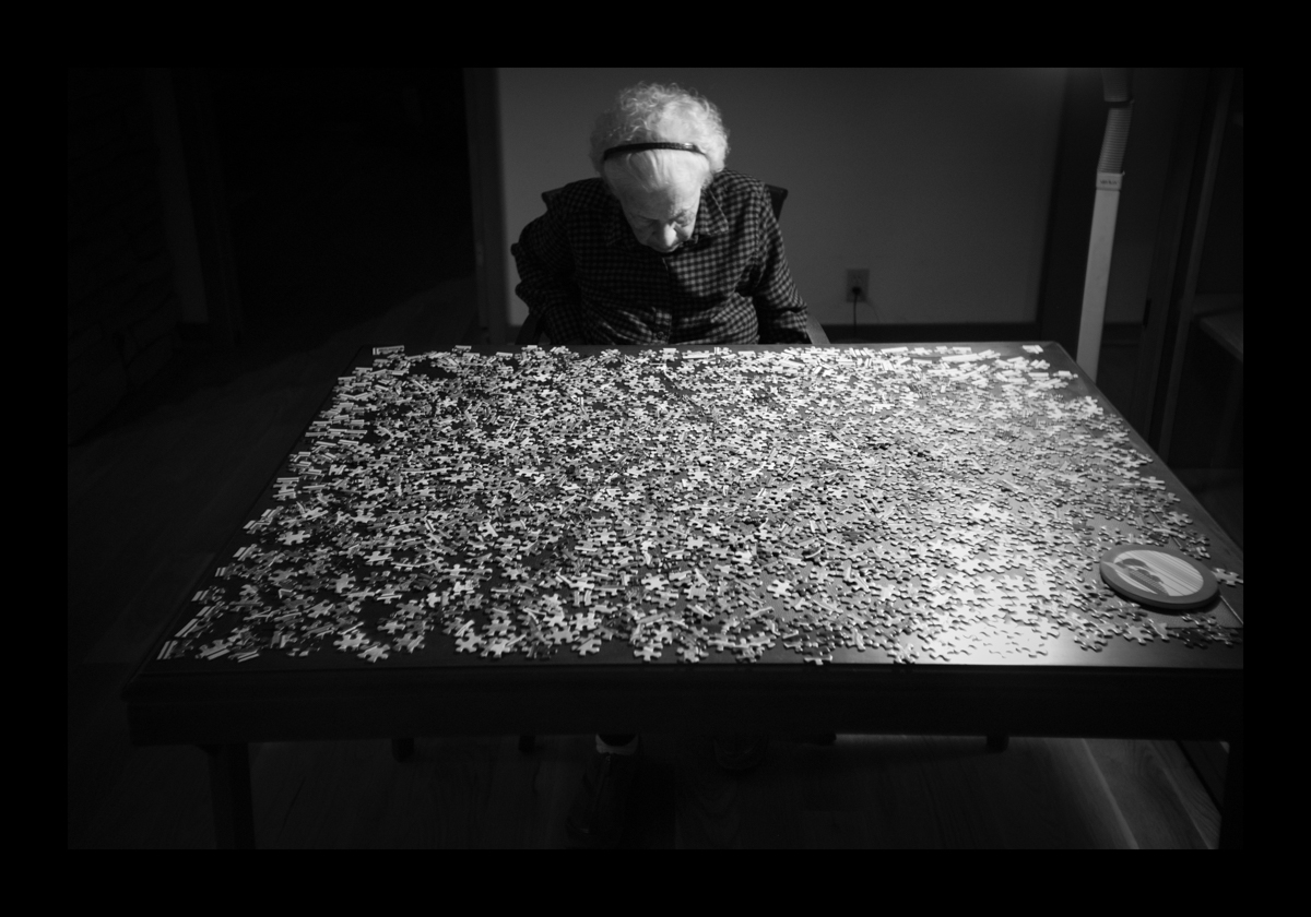 An older woman sits in front of a jigsaw puzzle unassembled on a table with her head down. © Virgil DiBiase, "1000 Pieces"