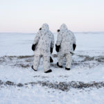 Two men walking across snow bundled in warm coats and hoods.