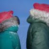 Two people stand back to back. Both are in green jackets with fur collars and a red hat.