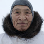 A portrait of an Inuit man with white coat, brown color and scull cap.