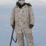 A man in white camouflage coat and pants against a backdrop of white snow. He holds a tool to break ice up into ice blocks..