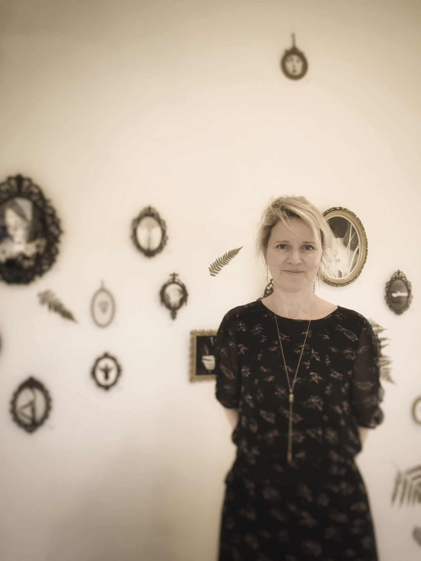 A woman stands in front of a wall full of small frames. She smiles graciously and has her arms around her back.